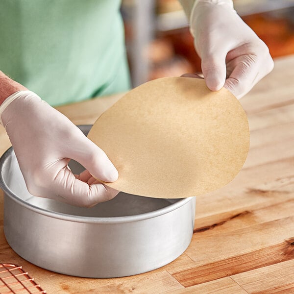 A gloved hand placing a round unbleached parchment liner in a round metal pan.