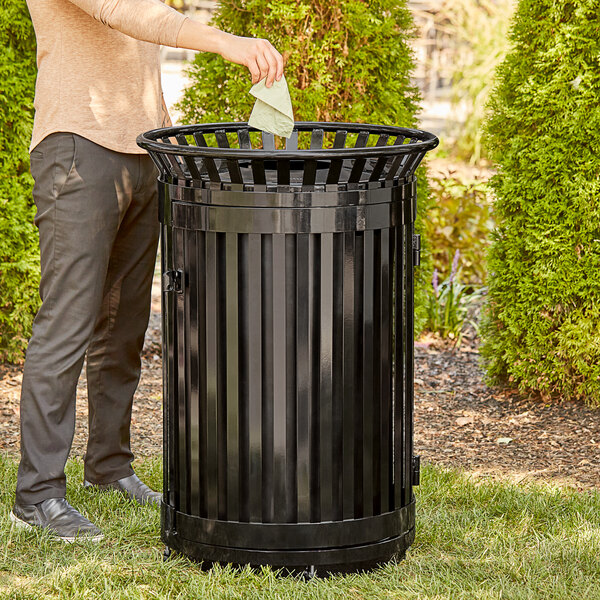 A person standing next to a Lancaster Table & Seating black steel outdoor trash can with a flat lid and door, putting a paper in it.