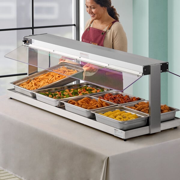 A woman wearing a red apron standing behind a stainless steel counter with a ServIt lighted heated buffet warmer full of food.