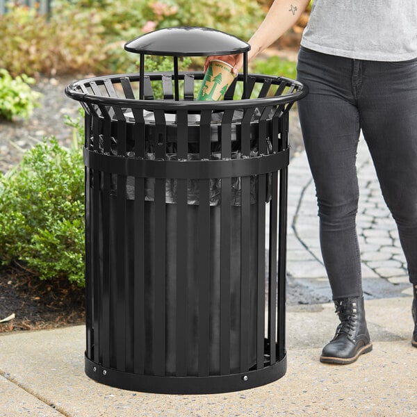 A woman standing next to a black LT&S round slatted steel trash can with a rain bonnet lid.