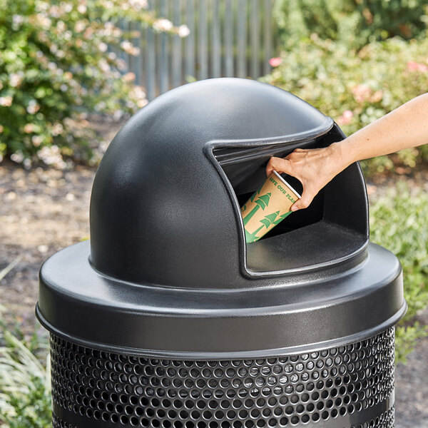 A hand using a Lancaster Table & Seating black plastic round dome lid to put a coffee cup in a trash can.