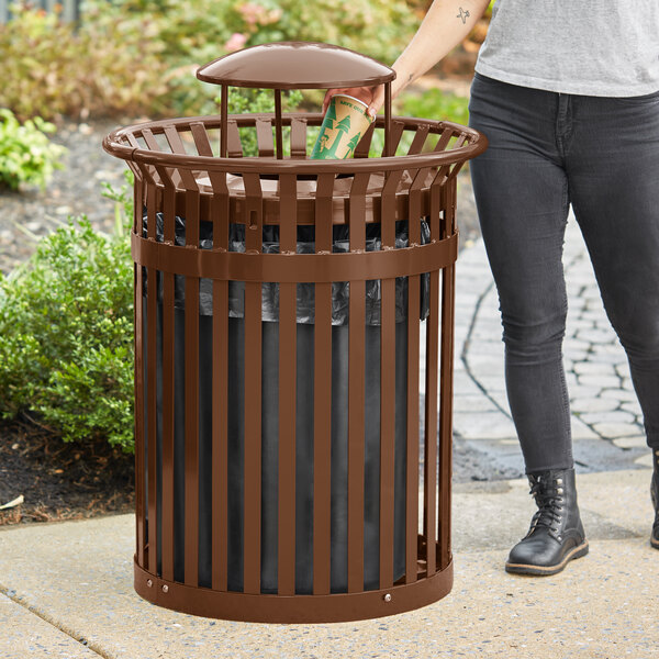 A woman standing next to a brown LT&S round slatted steel outdoor trash can with a black bag in it.