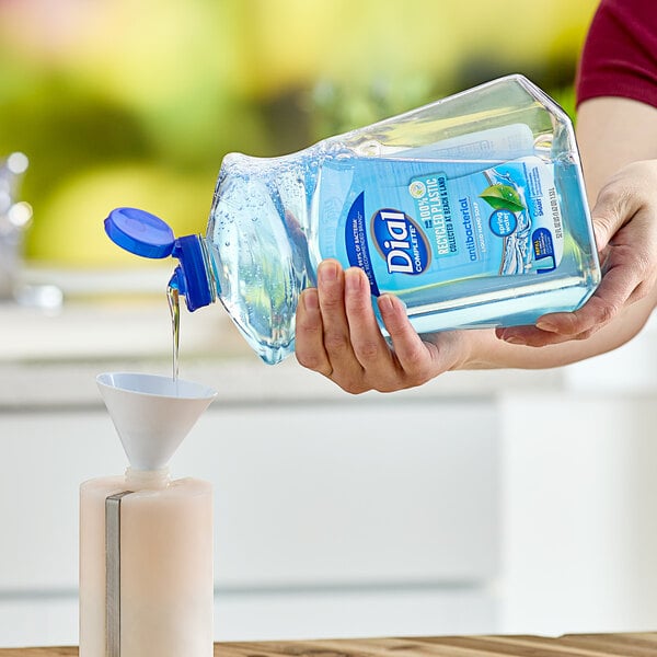 A person pouring Dial Complete Spring Water liquid hand soap into a plastic container using a white funnel.