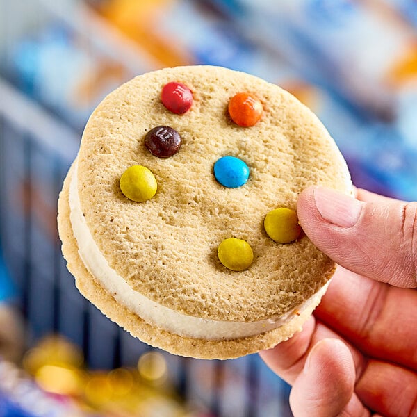 A hand holding an M&M's ice cream sandwich cookie with candy on it.