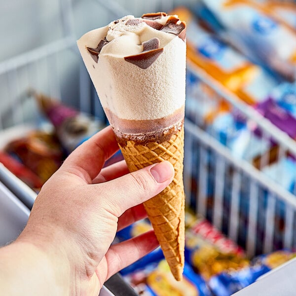 A hand holding a Blue Bunny brownie sundae frozen dessert cone with white and brown ice cream.