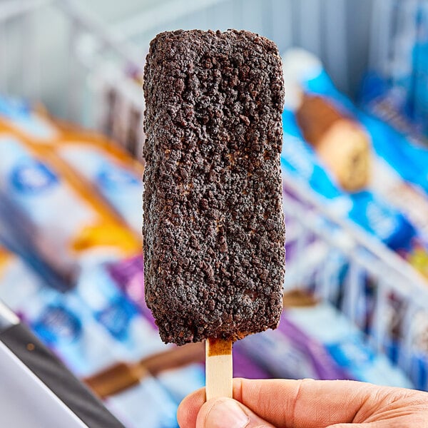 A hand holding a Blue Bunny Cookies 'n Cream frozen dessert bar with chocolate coating.