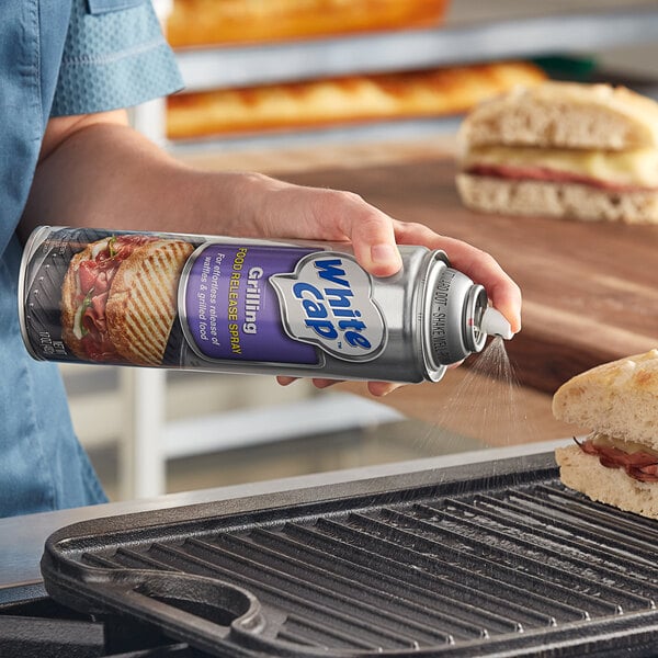 A person spraying a sandwich on a grill with White Cap Grill Release Spray.