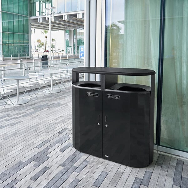 A black Rubbermaid outdoor waste receptacle on a brick patio.