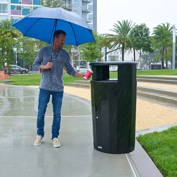 A man holding a blue and white umbrella standing next to a Rubbermaid outdoor waste receptacle.