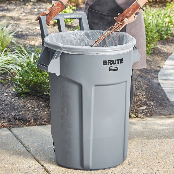 A woman putting a plastic bag into a Rubbermaid gray wheeled trash can.