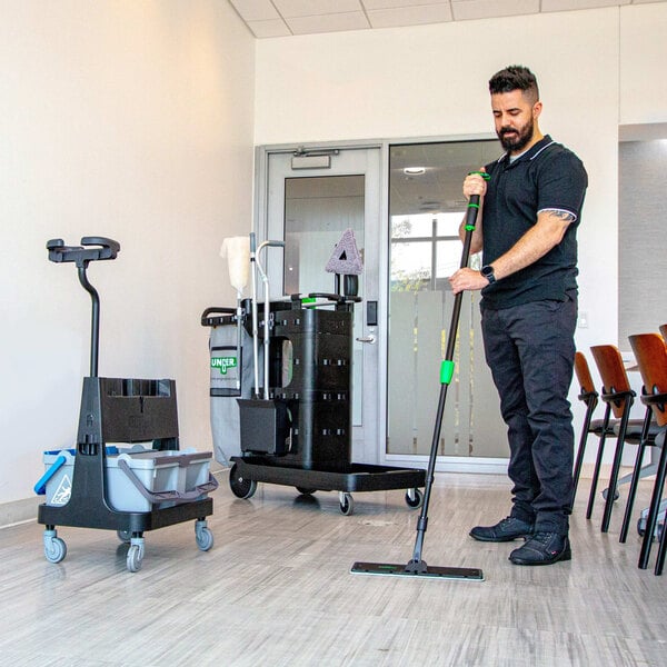 A man using a Unger Deep Cleaning Janitorial Cart System with a green and black handle mop to clean a room.