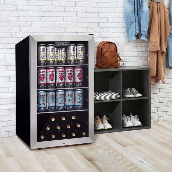 A stainless steel glass door beverage refrigerator filled with cans and bottles, placed next to a cubby shelf with shoes and a backpack.