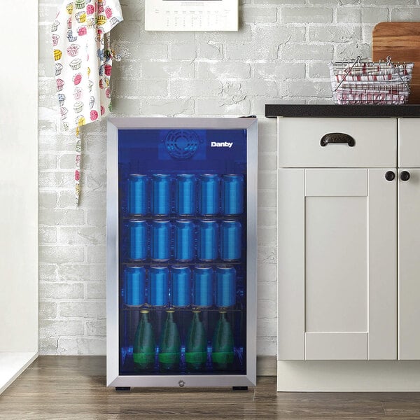 A stainless steel glass door beverage refrigerator filled with cans and bottles, placed next to a kitchen counter.