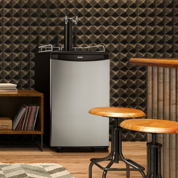 A Danby Spotless Steel double tap beer dispenser on a round table with round wooden stools.
