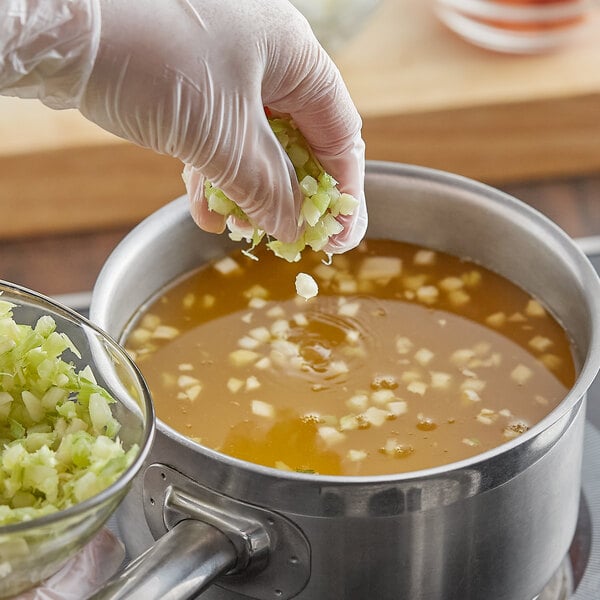 A gloved hand adds fresh diced celery to a pot of soup.