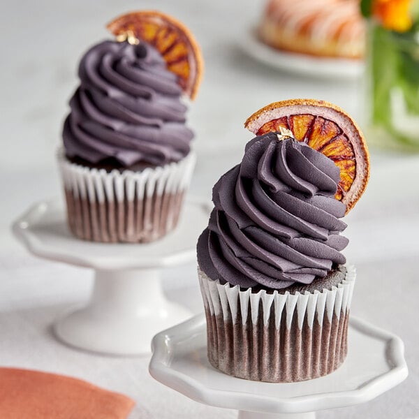 Two cupcakes with purple frosting and orange slices on top.