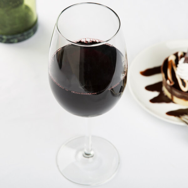 A Libbey Vina wine glass filled with red wine next to a plate of chocolate cake.