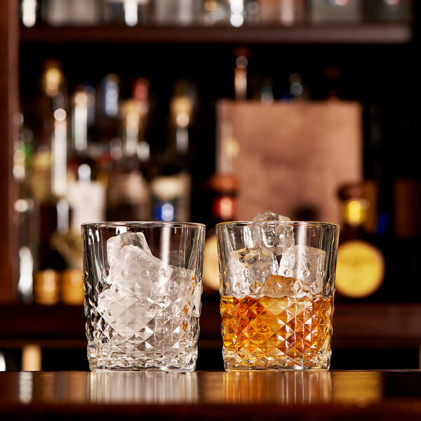 A pair of textured 12 oz. rocks glasses, one filled with ice and the other with an amber beverage and ice, displayed on a bar counter.