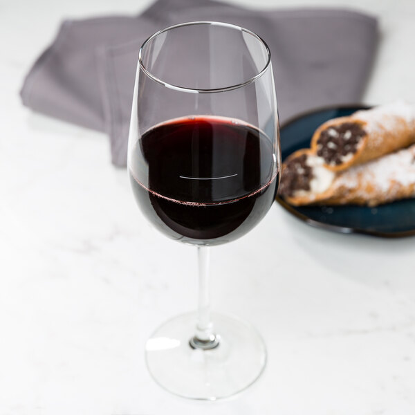 A Libbey tall wine glass filled with red wine next to a plate of pastries on a marble table.