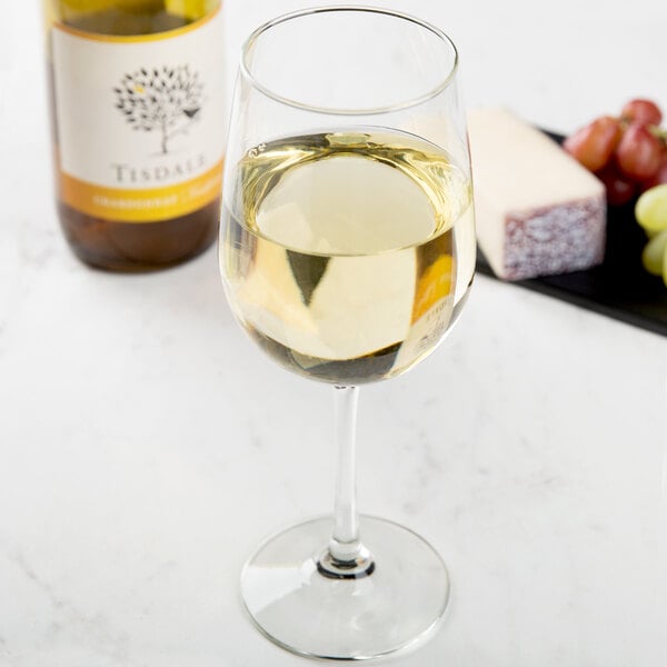 A Libbey Vina tall wine glass of white wine next to a bottle of wine and cheese.