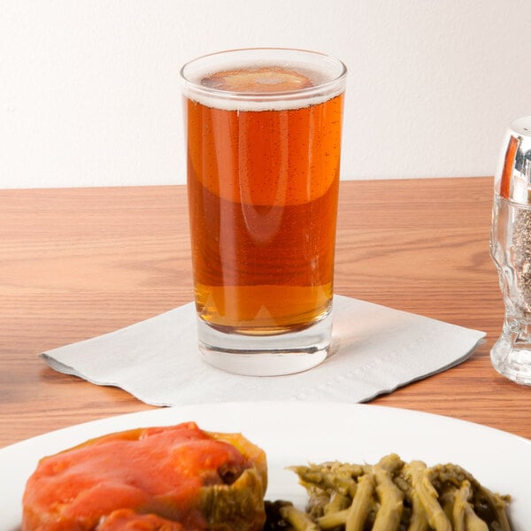 A Libbey highball glass filled with beer next to a plate of food on a table.