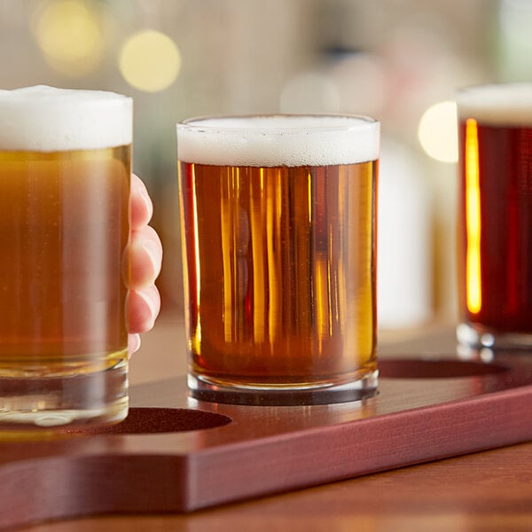 A hand holding a Libbey side water/tasting glass of beer on a coaster.