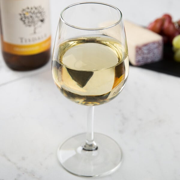 A Libbey Vina wine glass filled with white wine on a marble table with grapes and cheese.