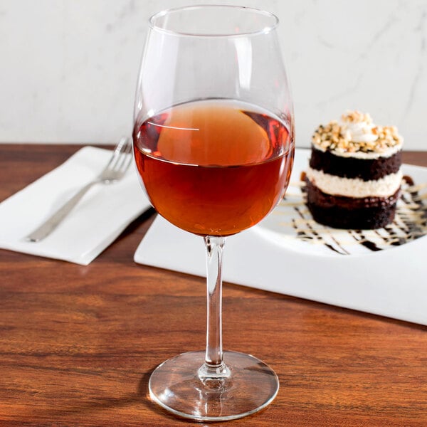 A Libbey wine glass filled with red wine next to a slice of cake on a table.