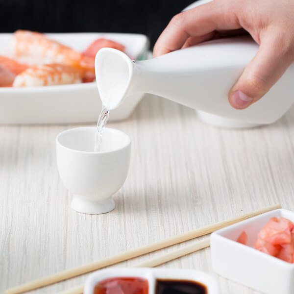 A person pouring white liquid into a small white 10 Strawberry Street sake cup.