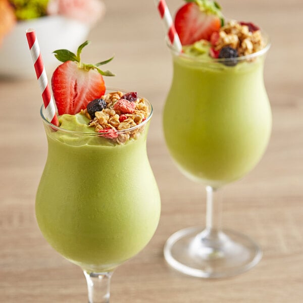 Two glasses of green smoothie with straws and fruit on a table.