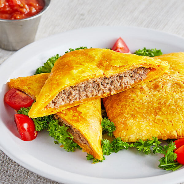 A plate of Caribbean Food Delights baked mild beef patties with a bowl of red sauce on a table.