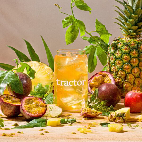 A glass of Tractor Beverage Co. Pineapple Passion Fruit juice next to a passion fruit and pineapple.