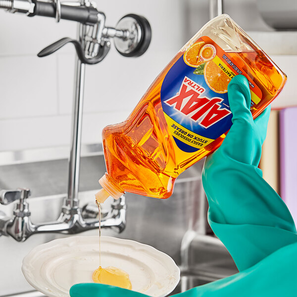 A person in gloves pouring Ajax Triple Action Orange Scent dish soap into a bowl.