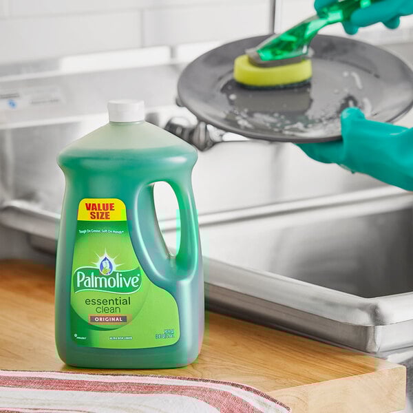 A person in green gloves using Palmolive Essential Clean dishwashing liquid to clean a pan in a home kitchen sink.