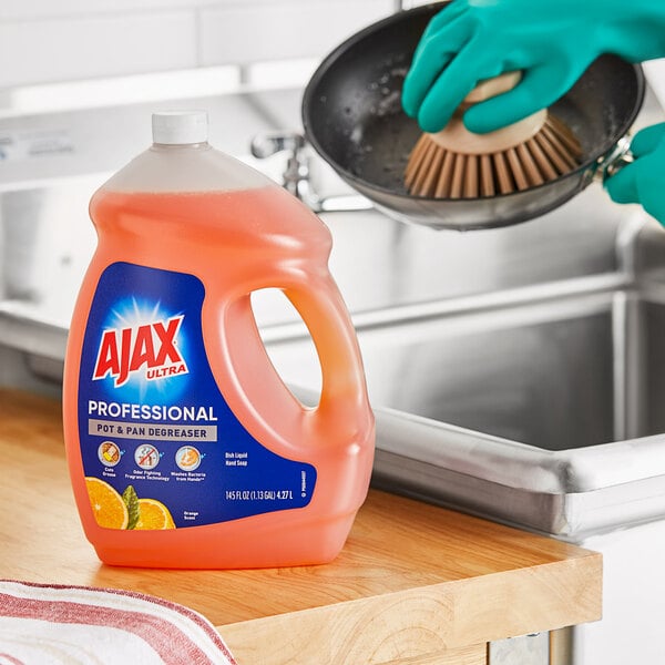 A gloved hand uses Ajax dish detergent to wash dishes in a sink.