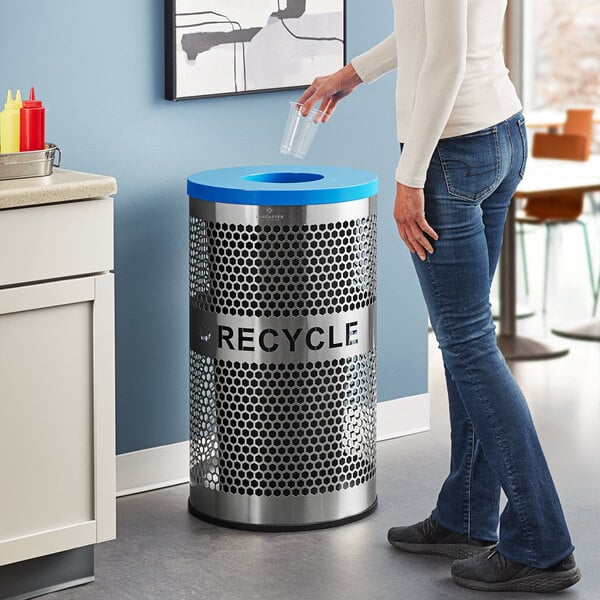 A woman in jeans throws a plastic cup into a Lancaster Table & Seating stainless steel recycling bin with a bottle lid.