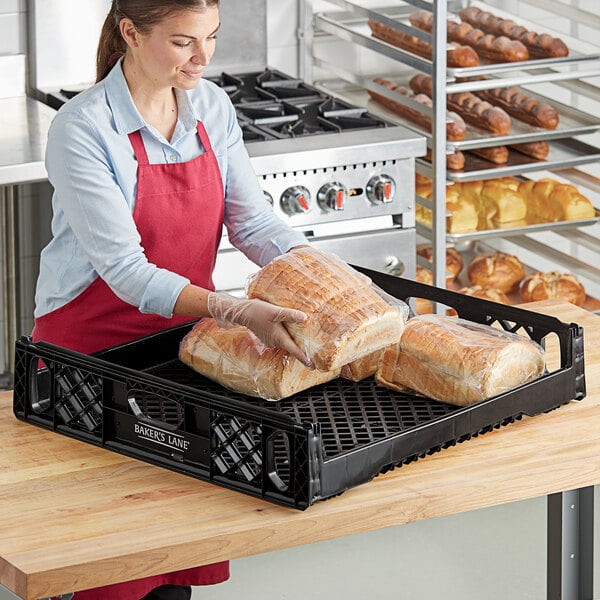 A woman in a red apron using a Baker's Lane black bread tray to display a loaf of bread wrapped in plastic.