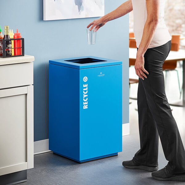 A woman pouring water into a blue Lancaster Table & Seating square decorative recycling bin.