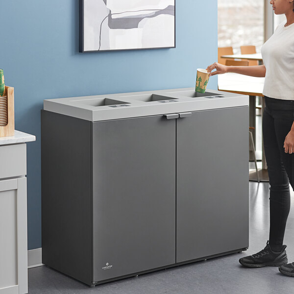 A woman standing next to a Lancaster Table & Seating dark gray steel rectangular three-stream waste receptacle.