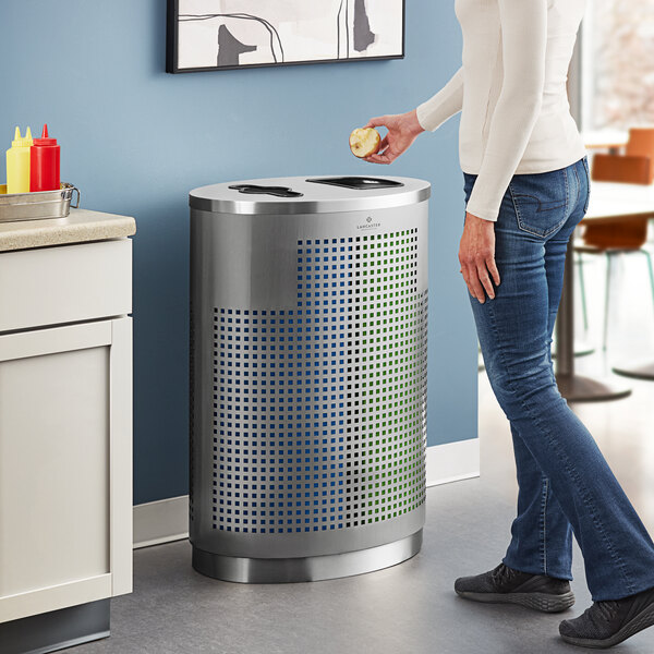A woman standing next to a Lancaster Table & Seating stainless steel trash can with trash and recycle openings.
