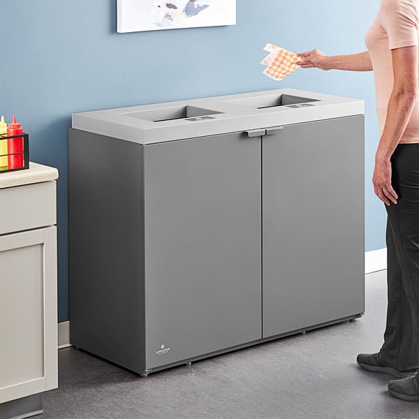 A woman standing next to a Lancaster Table & Seating dark gray rectangular two-stream waste receptacle.