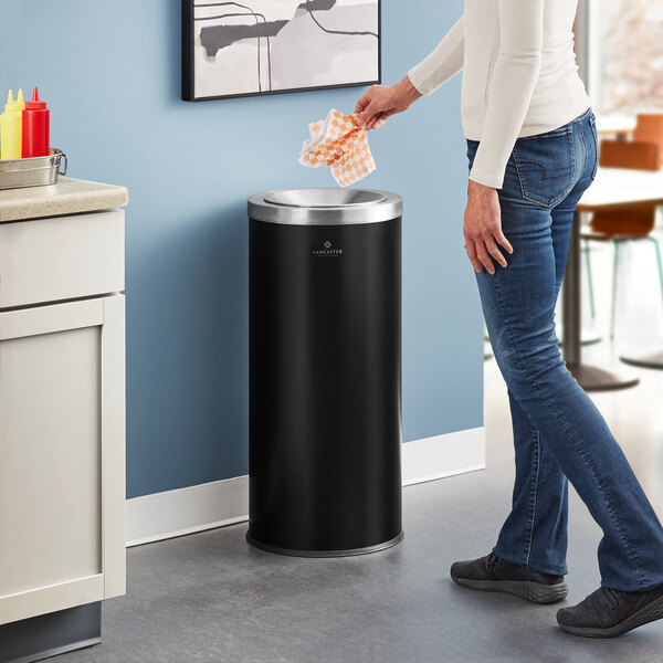 A woman in jeans throwing paper into a Lancaster Table & Seating black steel decorative trash can.