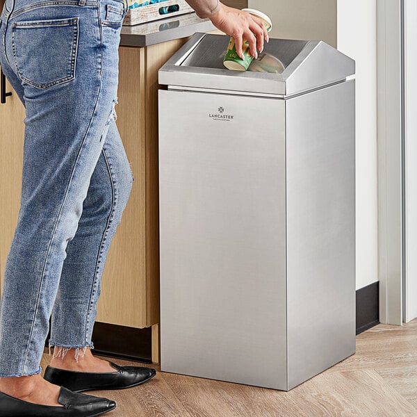 A woman in jeans uses a Lancaster Table & Seating stainless steel decorative trash can with a green and gold cup to throw something away.