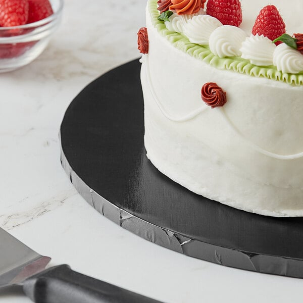 A close up of a Baker's Lane black cake drum with a cake frosted and topped with strawberries and raspberries.