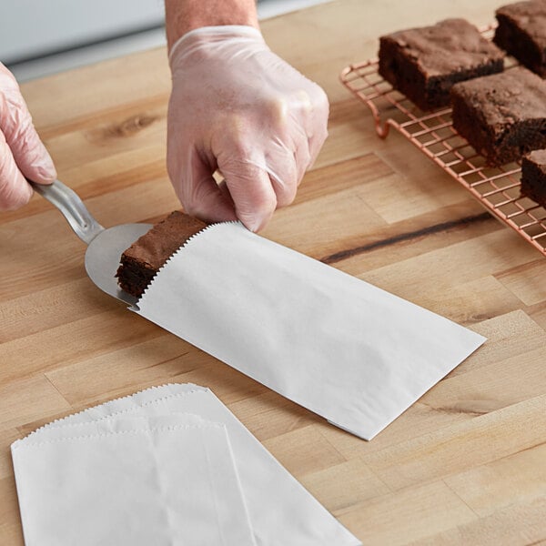 A hand using a spatula to put a brownie in a white paper bag.