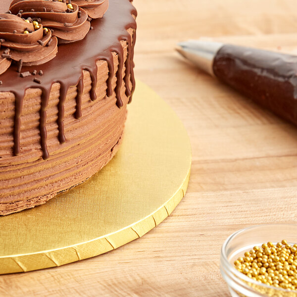 A chocolate cake with chocolate icing on a Baker's Lane gold cake drum on a table in a bakery display.