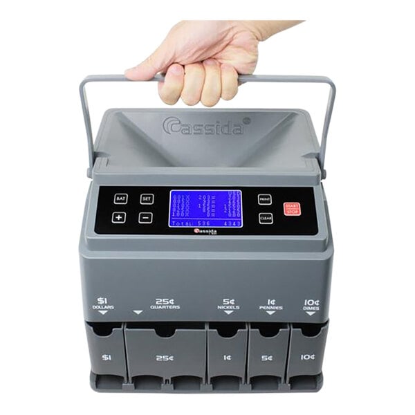 A hand holding a grey Cassida CC300 coin counter with a blue LED display.