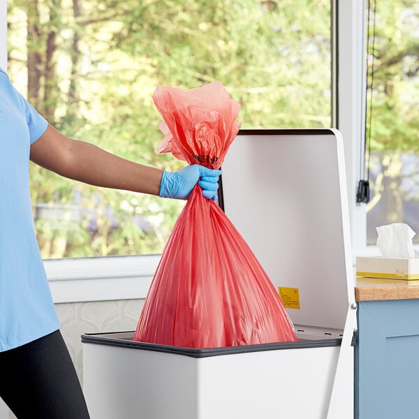 A person in blue gloves putting a red plastic bag into a trash can.