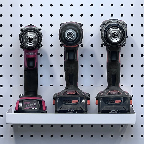 Three cordless drills on a white Triton shelf above a pegboard.