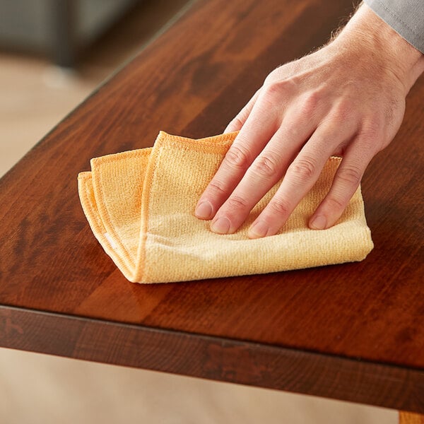 A hand wiping a wooden table with an orange Lavex microfiber cloth.